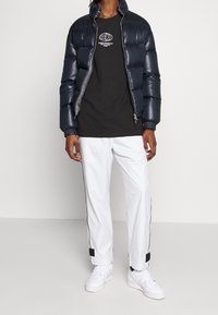 Granatowa kurtka puchowa z błyszczącą teksturą, czarny T-shirt z grafiką, białe spodni z wąskimi nogawkami z czarnym paskiem po boku oraz białe sneakersy.