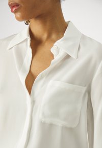 Hvid skjorte med krave, lavet af glat stof, med en brystlomme og en let glans. Lidt åben ved halsudskæringen.