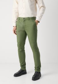 Zelene bombažne chinos hlače s slim fit krojem, zadrgo in stranskimi žepi, v kombinaciji s črnimi loaferji. Gladka tekstura in enotna barva.