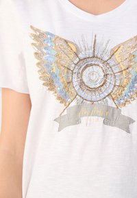 Bílý bavlněný tričko s dekorativní grafikou, která obsahuje barevné flitry a výšivku, včetně kruhového motivu a stuhy.