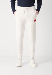 Pantalones deportivos blancos con cintura elástica y cordón, con un parche de logo rojo, piernas ajustadas y una textura lisa. Estilo casual.