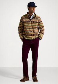 Többszínű pulóver geometriai mintákkal, zöld és barna árnyalatokban, bordó kordbársony nadrággal és barna bőr cipővel párosítva.