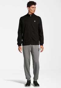 Giacca nera con zip e colletto alto, polsini e vita elasticizzati. Pantaloni tuta grigi con logo in evidenza. Scarpe sportive nere.