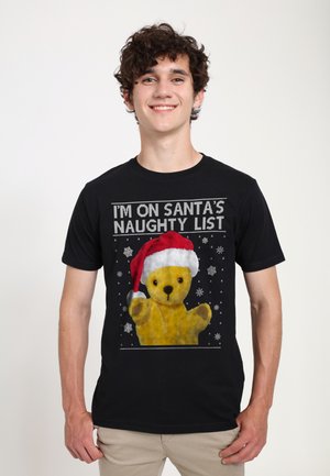 Sort T-shirt med et grafisk design af en gul teddy-bear iført en rød nissehue, med teksten "JEG ER PÅ SANTAS SLEMME LISTE" og snefnug.