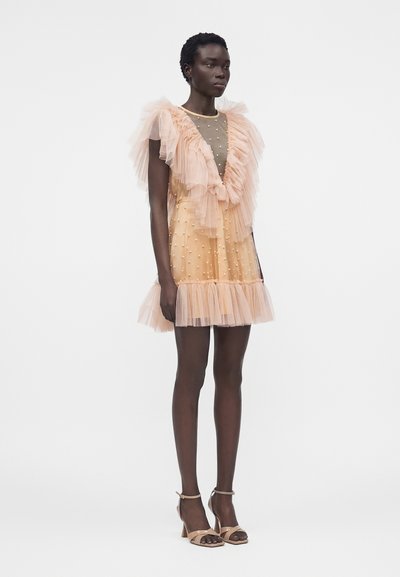 Γυναίκα με σκούρο δέρμα που φορά ένα ανοιχτό ροδακινί τούλινο φόρεμα με βολάν και μαργαριταρένιες λεπτομέρειες, σε συνδυασμό με γυμνά ψηλοτάκουνα πέδιλα, στέκεται μπροστά σε λευκό φόντο.