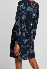 Blommönstrad klänning i mörk marinblå med gröna och rosa blad. Har långa ärmar, en åtsittande midja och en nyckelhålsdetalj på ryggen.