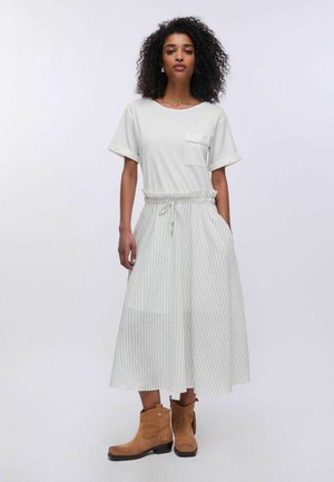 Donna con capelli ricci che indossa una maglietta bianca a maniche corte, gonna midi a righe con vita con coulisse e stivaletti alla caviglia in camoscio color marrone chiaro in piedi.