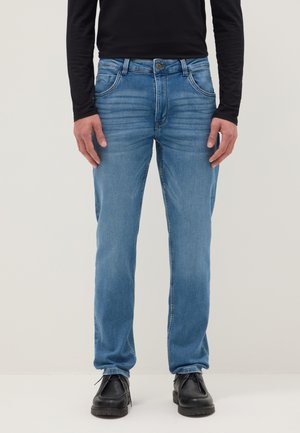 Hombre vestido con vaqueros de corte recto de color azul claro, camiseta de manga larga negra y zapatos de cuero negros, de pie frente a un fondo liso.