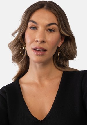 Woman with shoulder-length wavy hair wearing gold hoop earrings and a black V-neck top, looking directly at the camera with a neutral expression.