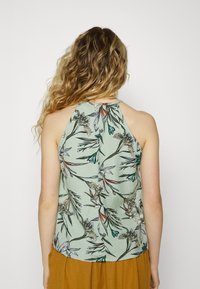 Top com padrão floral em tecido verde-claro, apresentando um corte descontraído, design costas abertas e detalhe de botões no pescoço.