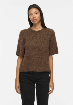 Femme portant un pull court marron duveteux et un pantalon noir taille haute, debout devant un fond uni.