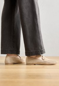 Beige semskede slip-on sko med flat såle, med en sydd detalj og minimalistisk design, kombinert med grå vide bukser.