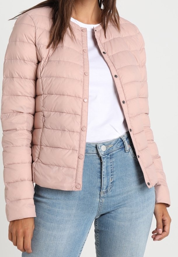 Femme portant une veste matelassée rose clair sur un t-shirt blanc et un jean bleu, debout devant un fond uni clair.