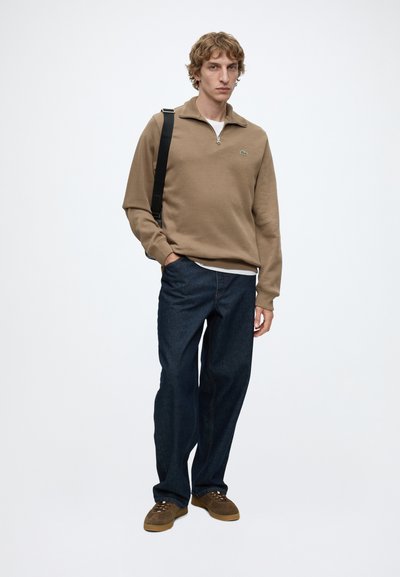 Jeune homme portant un pull zippé marron, un jean bleu foncé, des chaussures marron et un sac à bandoulière noir sur un fond blanc uni.