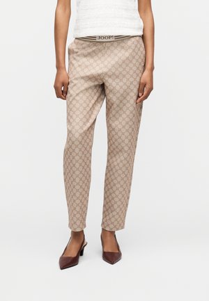 Persoană care poartă pantaloni bej cu model, top alb și pantofi cu toc maro, cu vârf ascuțit, stând în fața unui fundal alb simplu.