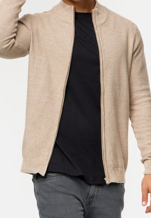 Homme portant un pull beige zippé sur un t-shirt noir et un pantalon gris, avec un bras levé près de la tête.