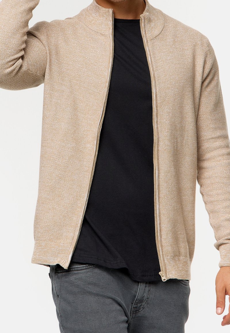 Homme portant un pull beige zippé sur un t-shirt noir et un pantalon gris, avec un bras levé près de la tête.