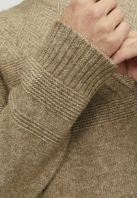 Maglione lavorato a maglia beige con motivi testurizzati, polsini a coste e una finitura morbida, che mostra cuciture dettagliate e spessori di tessuto variabili.