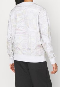 Bílá mikina s kulatým výstřihem, zdobená iridescentním vlnovým vzorem, s žebrovanými manžetami a spodním lemem. Vyrobena z měkkého materiálu, s uvolněným střihem a bočním švem.