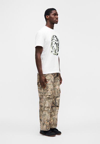 Λευκό βαμβακερό μπλουζάκι με γραφικό τύπωμα, συνδυασμένο με μπεζ cargo παντελόνια σε σχέδιο παραλλαγής και μαύρα παπούτσια με επίπεδη σόλα.