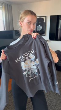 Νεαρή γυναίκα κρατάει μια σκούρα φούτερ με λευκό και χρυσό λουλουδένιο σχέδιο και την λέξη «BOTANICAL» τυπωμένη πάνω της.