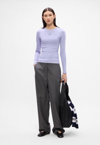 Lila Langarmoberteil mit gerippter Textur, graue gestreifte Hose, schwarze Ballerinas und eine schwarze Tasche mit einem weißen und blauen gemusterten Gegenstand.