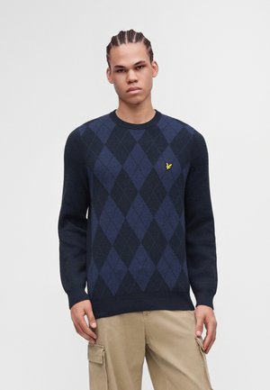 Ung mand med flettet hår iført en marineblå og sort argyle sweater og beige cargobukser mod en ensfarvet lys baggrund.