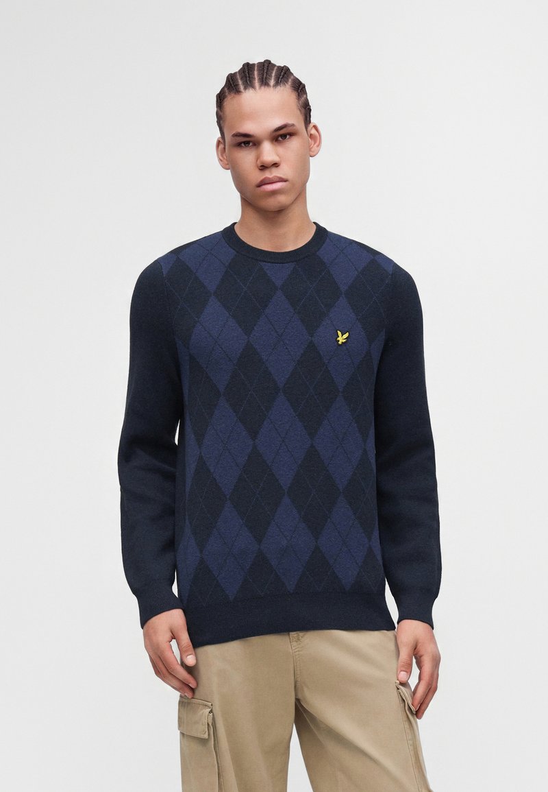 Jeune homme aux cheveux tressés portant un pull à losanges bleu marine et noir ainsi qu'un pantalon cargo beige sur un fond clair uni.