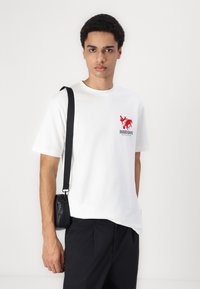 Hvid bomulds t-shirt med korte ærmer, der har et rødt trykt design på brystet. Sort crossbody taske med en rem hviler på skulderen.