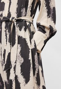 Μακρύ φόρεμα με σχέδιο που διαθέτει αφηρημένο εκτύπωμα σε μαύρο και μπεζ χρώμα, με ζώνη στη μέση, κουμπιά και υφή υφάσματος.