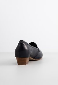 Chaussure en cuir noir avec un petit talon empilé, bout arrondi et une garniture décorative. Texture lisse et design simple.