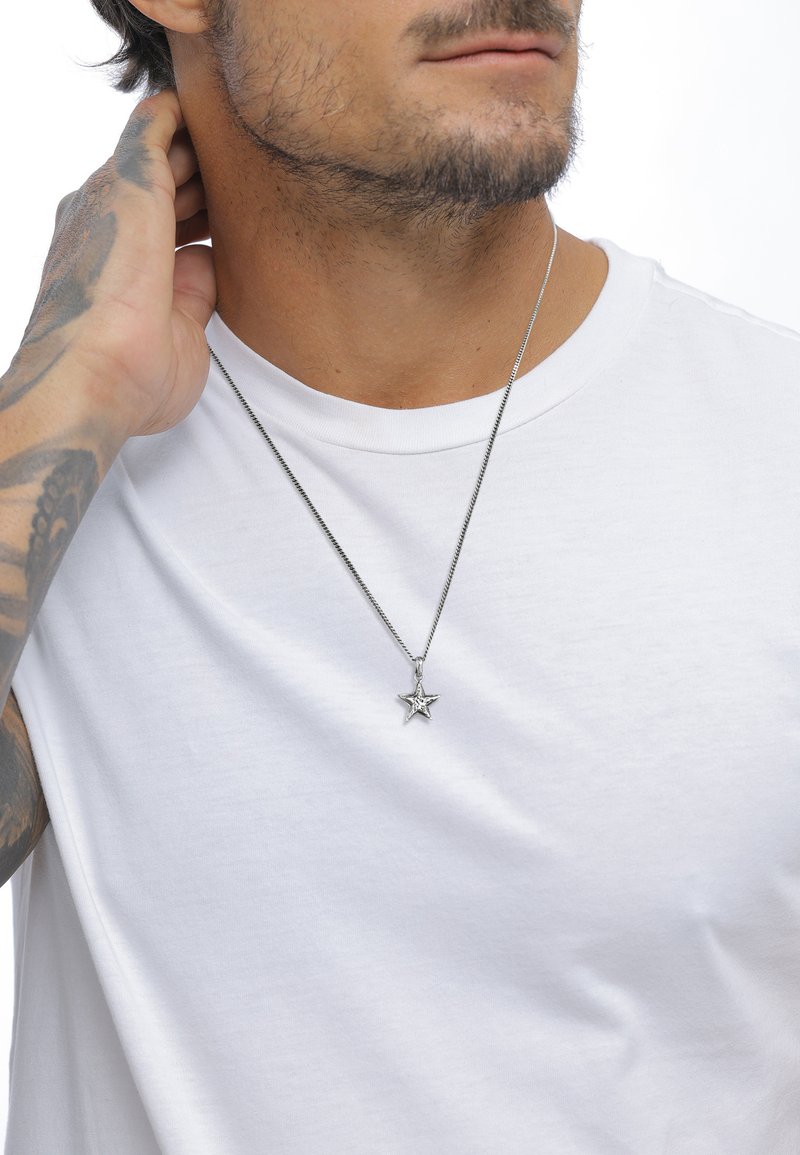 Silver star pendant necklace with a textured design, hanging from a thin metal chain, worn over a plain white t-shirt.