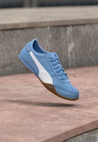 Baskets bleues avec une bande blanche et une semelle en gomme flottant au-dessus d'une surface en pierre avec un fond en béton.