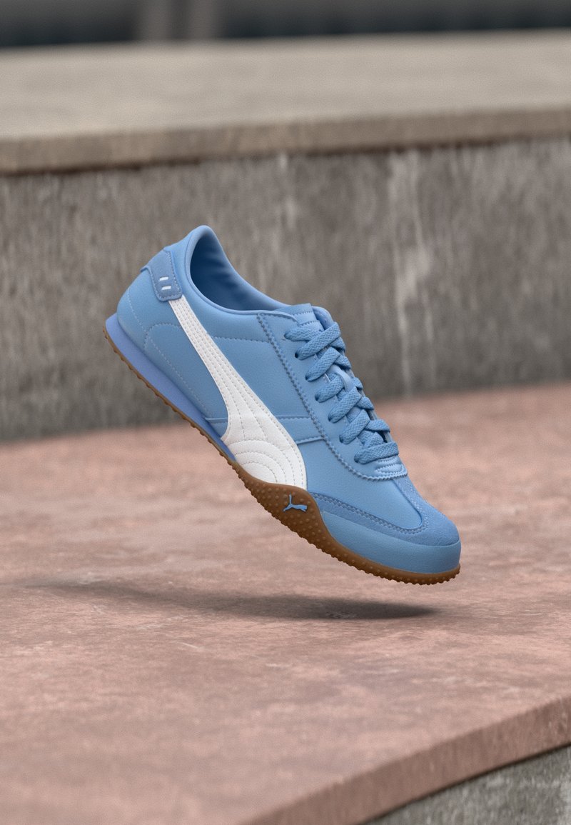 Baskets bleues avec une bande blanche et une semelle en gomme flottant au-dessus d'une surface en pierre avec un fond en béton.