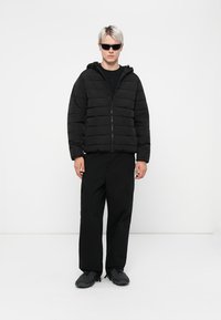 Veste rembourrée noire avec fermeture éclair et capuche, associée à un pantalon cargo noir et des chaussures noires. Modèle portant des lunettes de soleil, posé contre un fond blanc.