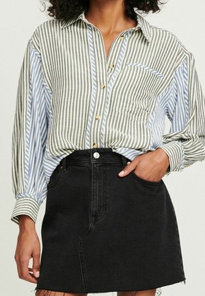 Kvinde iført oversize skjorte med vertikale striber i sort, hvid og blå samt sort denim mini nederdel, hånd i lommen.
