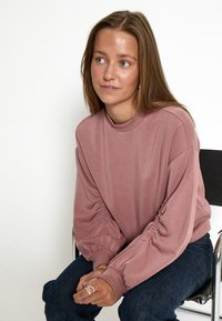 Prachový ružový top s dlhým rukávom a zhromaždenými rukávmi, s vysokým výstrihom, v kombinácii s tmavými džínsami. Hladká textúra, minimalistický dizajn.