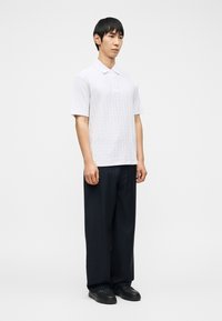 Άνδρας που στέκεται φορώντας λευκή πόλο με κοντά μανίκια και υφή, καθώς και χαλαρά μαύρα παντελόνια με λεπτές ρίγες και μαύρα παπούτσια, σε απλό φόντο.