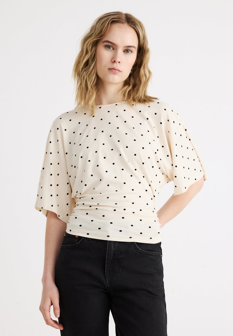 Γυναίκα με ώμο-μήκος κυματιστά ξανθά μαλλιά, ντυμένη με εκρού μπλούζα κοντομάνικη με μαύρες πουά και μαύρο παντελόνι, σε λευκό φόντο.