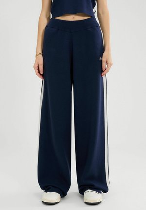 Kobieta w granatowych spodniach typu wide-leg z białymi paskami po bokach, białych sneakersach, krótkim granatowym topie, złotej bransoletce i pierścionku, stoi na białym tle.