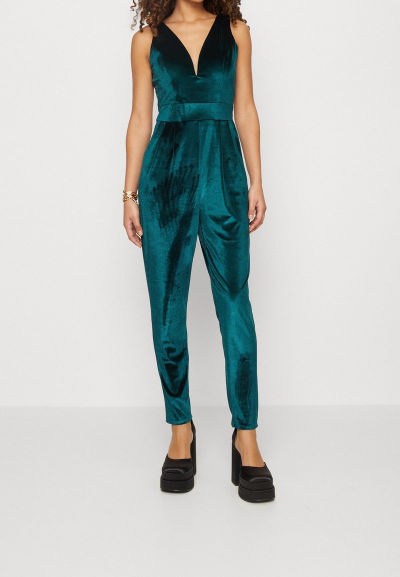 Teal sammetsbyxdress med djup V-ringning, figursydd midja och avsmalnande ben. Har en mjuk textur och minimalistisk design, matchat med svarta platåsandaler.
