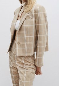 Traje de cuadros beige con un blazer ajustado y pantalones a juego. La tela tiene una textura suave, con delgadas líneas blancas y un detalle en el bolsillo delantero.