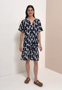 Femme debout portant une robe mi-longue avec un motif abstrait bleu marine et blanc et des chaussures marron à enfiler dans une pièce neutre.