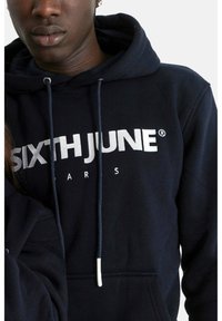 Mörkblå hoodie gjord av mjuk bomull, med vit broderad logotyp "SIXTH JUNE PARIS." Inkluderar dragsnören och framsida ficka.