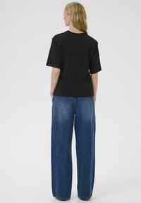 Modella femminile in piedi con la schiena rivolta alla fotocamera, indossa una camicia nera oversize e jeans blu a gamba larga su uno sfondo bianco semplice.