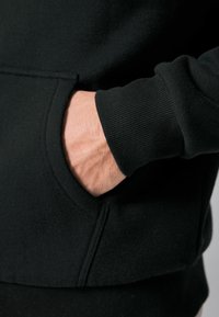 Sort hættetrøje med en frontlomme, ribbede manchetter og en blød tekstur, der viser en hånd hvilende i lommen.