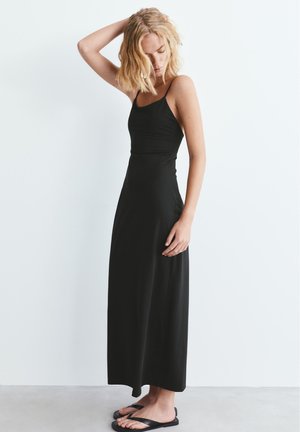 Blondínka v dlhých čiernych šatách na tenké ramienka a čiernych sandáloch stojaca pred jednoduchým bielym pozadím, dotýkajúca sa svojich vlasov.