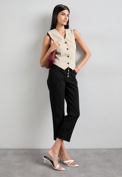 Gilet beige senza maniche con cinque bottoni neri, abbinato a pantaloni neri con dettagli a bottoni dorati. Tacchi bianchi aperti e una borsa bordeaux.