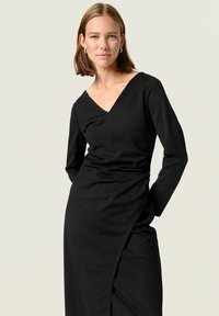 Vestido negro de manga larga con escote en V, que presenta un corsé ajustado y una falda drapeada, confeccionado en una tela suave y lisa.