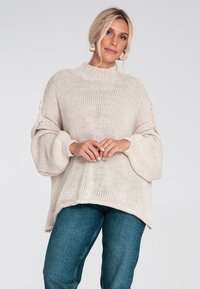 Beżowy dziany sweter z wysokim kołnierzem, oversized rękawami i prążkowanymi mankietami. Połączony z dopasowanymi niebieskimi dżinsami, minimalne detale tekstury.